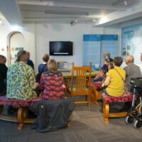 Group of participants, pictured from behind, sat down and watching a display on a television.