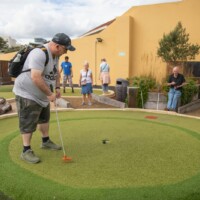 Group of participants playing outdoor crazy golf. Kev has just hit the ball and it's about to go into the hole.