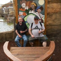 Two participants at an outdoor crazy golf place sitting in a prop boat and holding golf clubs.