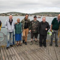 Group of particiapants standing with their backs against the railing and facing the camera. There's the ocean with boats in the background.