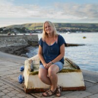 Rachel sat on an upturned boat on a stone pathway in front of the ocean.