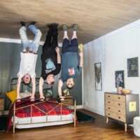 Three participants posing in the 'Upside Down House'. They're pretending to be standing on the ceiling, gripping a beds railing.