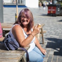 Gill sitting down on a bench at the seafront, smiling and eating an ice cream.