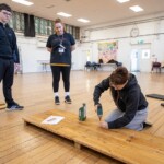 A young person using a drill to screw a plank of wood to another piece. One other young person is watching, as well as a supervisor.