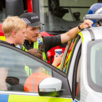 Some kids looking inside a police car, with one sitting in the driving seat. A police man is talking to them.